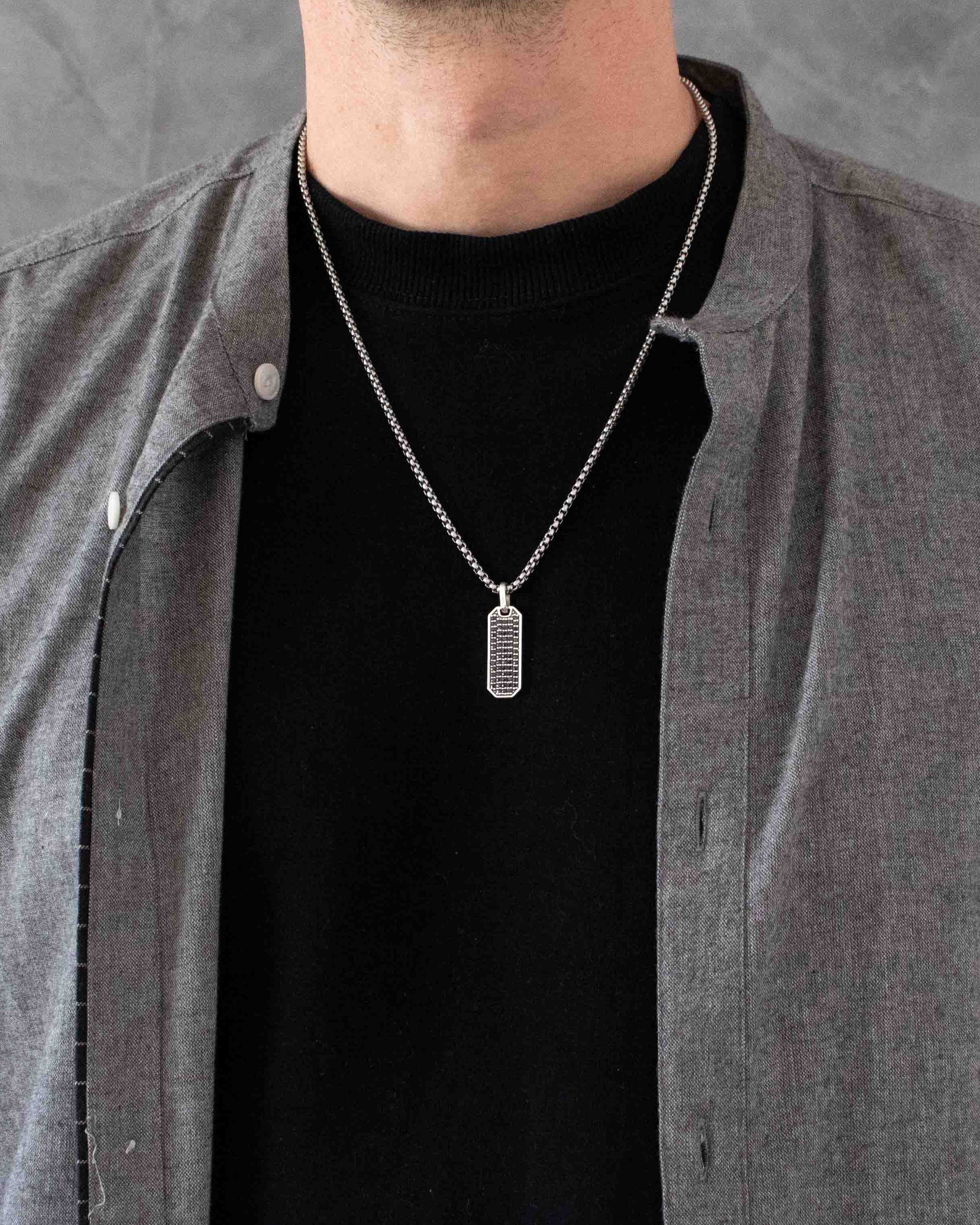 Man wearing a silver necklace with a rectangular pendant against a gray background